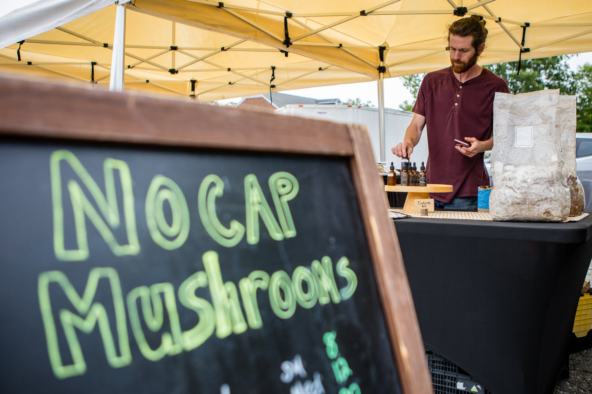 Founder, Nick,  standing behind a food stand with a sign reading 'No Cap Mushrooms' in an outdoor setting.